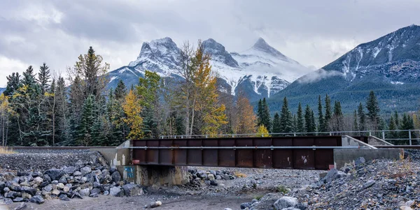 Sonbahar mevsiminde demiryolu manzarası, beyaz bulutlar ve kar kaplı mavi gökyüzü arka planda üç kız kardeş zirvesi üçlüsü. Canmore, Alberta, Kanada. — Stok fotoğraf