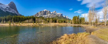Quarry Lake Park göl kıyısı sonbahar sezonunun sonlarında güneşli bir sabah. Arkaplanda mavi gökyüzü ile Rundle Dağı ve Lawrence Dağı Grassi Ha Ling Tepesi karla kaplı. Canmore, Alberta, Kanada.