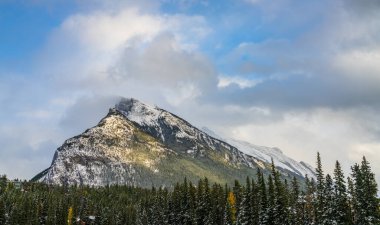 Karla kaplı Rundle Dağı sıradağları. Mavi gökyüzü üzerinde karlı orman ve kışın güneşli bir günde beyaz bulutlar. Banff Ulusal Parkı, Kanada Kayalıkları.