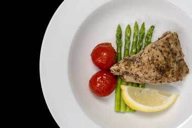 Yakın plan, kırmızı balık bifteği, kuşkonmaz, domates ve limon siyah arka planda beyaz bir tabakta. Tablo üstü görünümü.