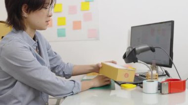 Asyalı kadın ofis çalışanı paket kutularını barkod etiketlerini okuyarak bilgisayardaki bilgileri kaydetmek için barkod tarayıcısıyla kontrol ediyor..
