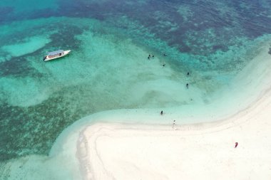 Mercan kayalıklarının üzerinde tekneyle mavi okyanus suyuyla yüzen ve beyaz kumsalda yürüyen insanların hava aracı görüntüsü. Tinggi Adası veya Pulau Tinggi, Malezya