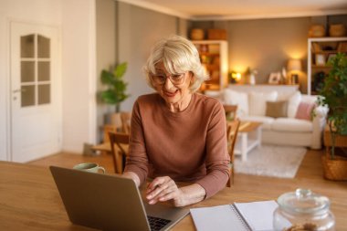 Modern oturma odasındaki masada otururken online terapi videosu için dizüstü bilgisayar kullanan neşeli son sınıf kadını.