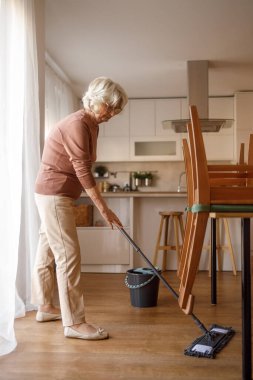 Yemek odasını temizlerken ahşap yerleri paspaslayan ve ev işlerini yapan yaşlı bir kadının yan görüntüsü.