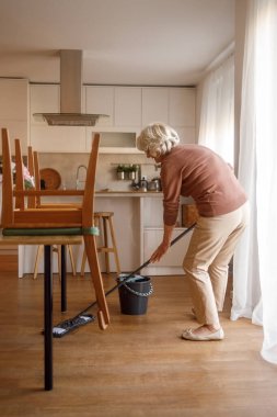 Yaşlı bir kadın, günlük ev işlerini yaparken paspas ve kova kullanarak parke mutfak zeminini paspaslıyor.