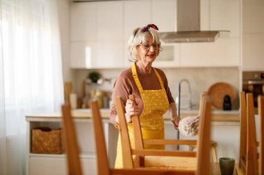 Önlüklü yaşlı kadın yemek odasında tozlu tahta sandalyeleri temizlerken evde ev işleri yapıyor.