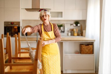 Önlük giymiş neşeli olgun bir kadının portresi. Evde toz beziyle yemek odası sandalyelerini ilaçlarken şarkı söylüyor.