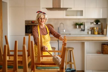 Önlük giyen olgun bir kadın modern mutfakta tozlu ahşap yemek sandalyelerinin tozunu ve kirini temizliyor.