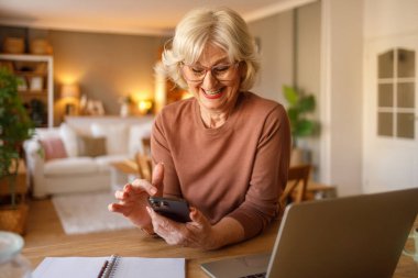 Masasında dizüstü bilgisayarı olan, gözlüklü neşeli yaşlı kadın evden alışveriş için cep telefonu kullanıyor.