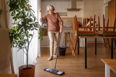 Kulaklıklı yaşlı kadın evde yemek masasının yanındaki ahşap zemini paspaslarken neşeyle dans ediyor.