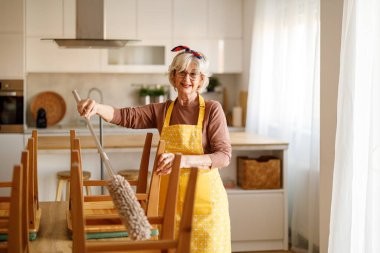 Önlüklü, kirli ahşap sandalyelerin tozunu alırken yemek odasında ev işlerini yaparken mutlu bir son sınıf kadını.