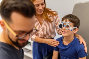 Bakıcı anne göz muayenesi sırasında oğluna bakıyor. Erkek doktor göz hekimliği kliniğindeki görüşünü kontrol ediyor.