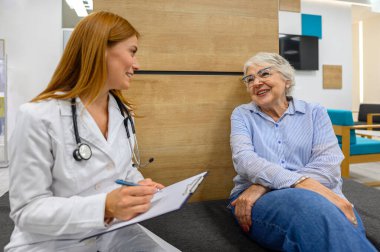 Klinikte sağlık sorunlarını anlatırken gülümseyen son sınıf öğrencisi kadın doktor notlar yazıyor.