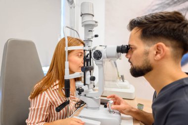 Erkek doktor göz hekimliği kliniğinde göz muayenesi sırasında yarık lamba ışığı kullanarak genç bir kadının gözlerini inceliyor.