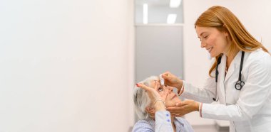 Kadın göz doktorundan göz damlası alan yaşlı bir kadın hastanedeki görme kontrolü sırasında beyaz duvara çarpıyor.