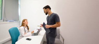 Erkek doktor elinde dosya tutuyor. Kadın meslektaşına danışıyor. Klinik masasındaki hasta kayıtlarını inceliyor.