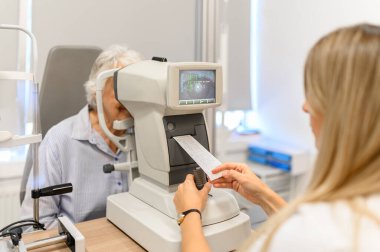 Klinikteki görme testi sırasında yaşlı kadının gözlerini incelerken kesik lambadan çıkan kâğıt çıktılarını inceleyen kadın göz doktoru.