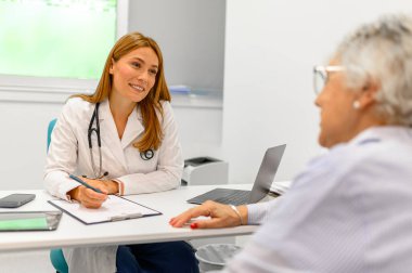 Yaşlı kadın muayenede semptomları ve tıbbi geçmişi anlatırken, kadın doktor da not alıyor.