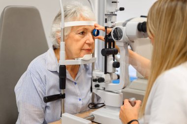 Yaşlı bir kadının gözlerini kesik lambayla inceleyen kadın göz doktoru klinikte teşhis lenslerini tutuyor.