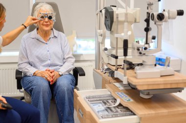 Yaşlı kadın göz hekimliği kliniğinde görme testine girerken, phoropter lenslerini ayarlayan kadın göz doktoru