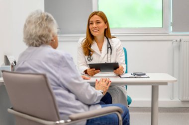 Hasta kayıtlarını tutan gülümseyen kadın doktor ve klinikteki sağlık sorunlarını anlatan yaşlı kadını dinliyordum.