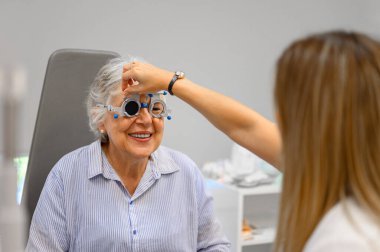Yaşlı bir kadın klinikte göz muayenesinden geçerken, phoropter lenslerini ayarlayan kadın göz doktoru.