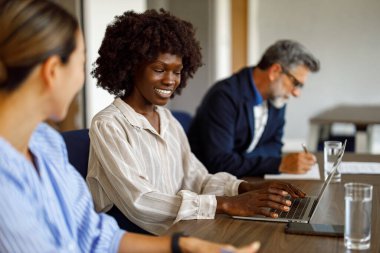 Bilgisayarda çalışırken iş arkadaşlarıyla tartışan ve modern yönetim kurulunda iş adamlarının yanında oturan mutlu iş kadını.