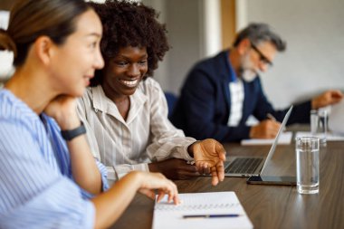 Mutlu bayan meslektaşlar iş stratejilerini görüşüyorlar. Laptop ve not defteri kullanıyorlar. Ofisteki iş adamlarıyla.