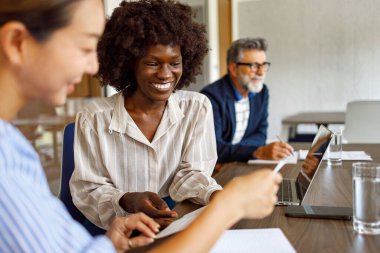 Gülümseyen kadın profesyoneller finansal belgeleri incelerken iş adamları modern ofiste işbirliği yapıyor.