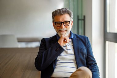 Toplantı masasında otururken kameraya gülümseyen, gözlüklü kendine güvenen erkek girişimcinin portresi.