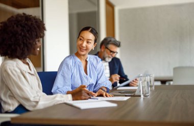 Kadın muhasebeciler mali raporları incelerken, erkek çalışanlar da kurumsal ofiste arka planda çalışıyor.