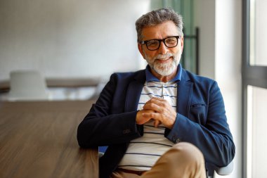 Gözlüklü, kameraya gülümseyen, güvenli bir şekilde toplantı odasında oturan sakallı erkek portresi.