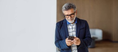 Gözlüklü olgun bir işadamı, şirket ofisinde beyaz duvarın yanında dururken akıllı telefondan mesajlaşır.