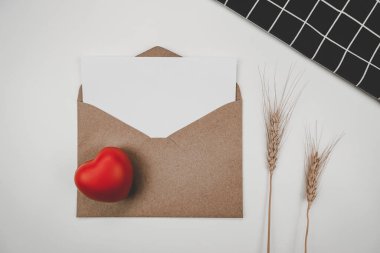 Boş beyaz kağıt, açık kahverengi bir zarfa, kırmızı bir kalbe ve arpa kuru çiçeğe ve beyaz arka planda siyah bir beze yerleştirilir. Sevgililer Günü konsepti