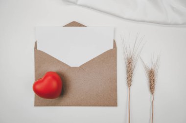 Boş beyaz kağıt, açık kahverengi bir zarfa, kırmızı bir kalbe, arpa kuru bir çiçeğe ve beyaz bir örtüye konur. Sevgililer Günü konsepti