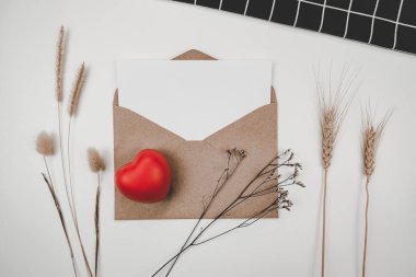 Boş beyaz kağıt, açık kahverengi bir zarfa, kırmızı bir kalbe, bir sürü kurutulmuş çiçeğe, beyaz arka planda siyah bir beze yerleştirilir. Sevgililer Günü konsepti