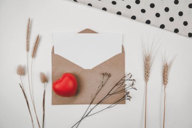 Boş beyaz kağıt, açık kahverengi bir zarfa, kırmızı bir kalple, bir sürü kurutulmuş çiçekle, beyaz arka planda beyaz bir bezle konur. Sevgililer Günü konsepti