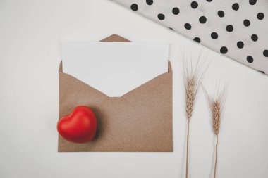 Boş beyaz kağıt, açık kahverengi bir zarfa, kırmızı bir kalbe, arpa kuru bir çiçeğe ve beyaz bir örtüye konur. Sevgililer Günü konsepti
