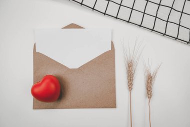 Boş beyaz kağıt, açık kahverengi bir zarfa, kırmızı bir kalbe, arpa kuru bir çiçeğe ve beyaz bir örtüye konur. Sevgililer Günü konsepti