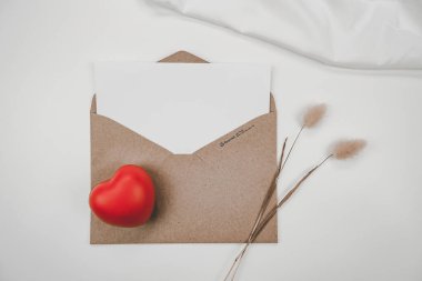 Boş beyaz kağıt, açık kahverengi kağıt zarfın üzerine yerleştirilir. Kırmızı kalp ve Tavşan kuyruğu kuru çiçek ve beyaz arka planda beyaz bez. Sevgililer Günü konsepti