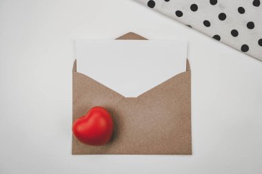 Boş beyaz kağıt, açık kahverengi bir zarfa, kırmızı bir kalbe ve beyaz bir arkaplanda beyaz bir beze yerleştirilir. Sevgililer Günü konsepti
