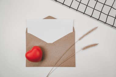 Boş beyaz kağıt, açık kahverengi bir zarfın üzerine yerleştirilir. Kırmızı kalp, kıvırcık kuyruklu kuru çiçek, beyaz arka planda beyaz bez. Sevgililer Günü konsepti