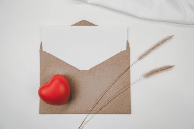 Boş beyaz kağıt, açık kahverengi bir zarfın üzerine yerleştirilir. Kırmızı kalp, kıvırcık kuyruklu kuru çiçek, beyaz arka planda beyaz bez. Sevgililer Günü konsepti