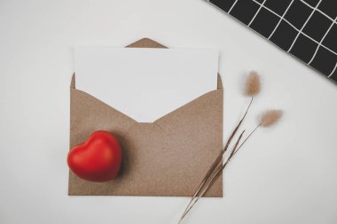 Blank white paper is placed on open brown paper envelope with red heart, Rabbit tail dry flower and black cloth  on white background. Valentine's day concept