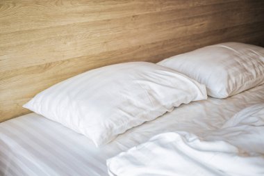 Beyaz yataklı yatak odası, beyaz iki yastık, beyaz yorgan, tahtadan yatak başlığı ve güneş ışığı olan yatak odası.