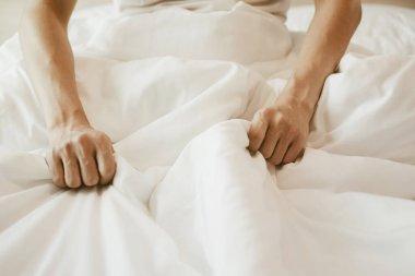 Yatakta oturan ve elinde beyaz bir battaniye tutan bir adam. Beyaz yataklı ve güneşli yatak odası..