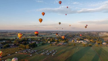 A vibrant display of hot air balloons fills the sky above a picturesque countryside at dawn. The breathtaking view showcases fields and small towns below, creating a serene atmosphere.