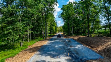 A construction crew works on a new road surrounded by tall trees and greenery in a rural setting. The sun shines brightly in the blue sky, highlighting the unpaved ground and their progress.