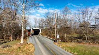 Etrafı yapraksız ağaçlarla çevrili dolambaçlı bir yolda kırsal bir köprü manzarası. Parlak mavi gökyüzü, ilkbaharın başlarındaki topraksı tonlara katıksız bir tezat sağlar..