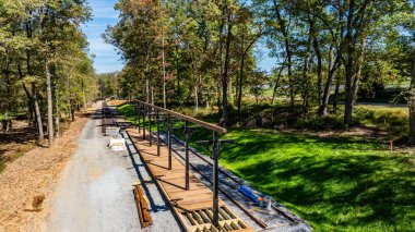 Builders are constructing a wooden platform alongside a gravel road in a wooded area. Surrounding trees provide a serene backdrop on a clear, sunny day, enhancing the tranquil environment.